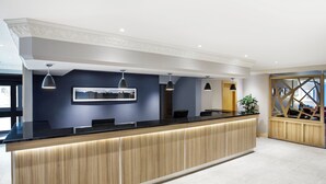 A reception desk with a wooden finish and a black countertop, flanked by two pendant lights and a framed picture above. - Leonardo Hotel Inverness (Inverness)