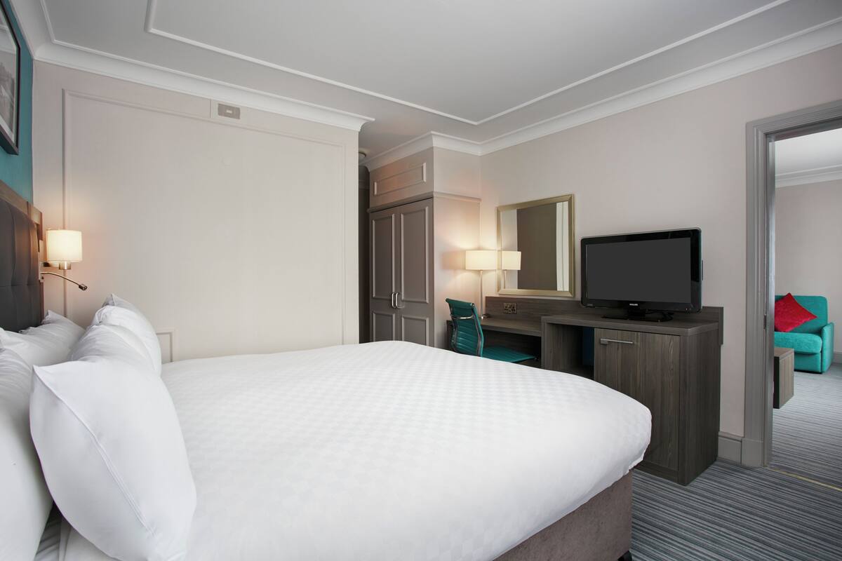 A hotel room with a large bed, a desk with a television, a chair, and a small seating area with a red pillow.