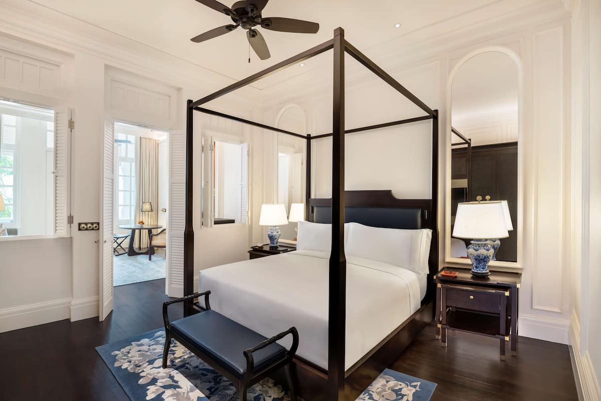 A four-poster bed with a ceiling fan, a bedside table with a lamp, and a view of a dining area through an open door.
