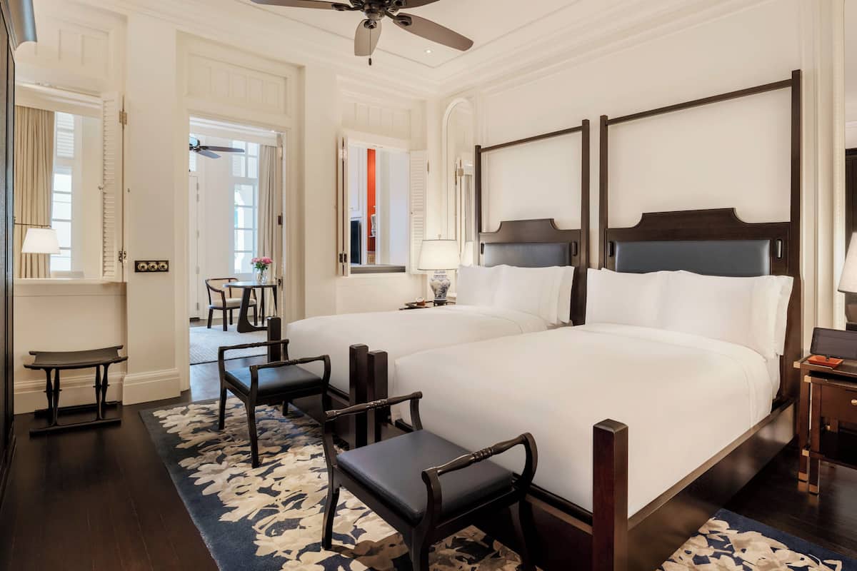 A hotel room with two beds, a ceiling fan, a desk with a chair, and a window with curtains.