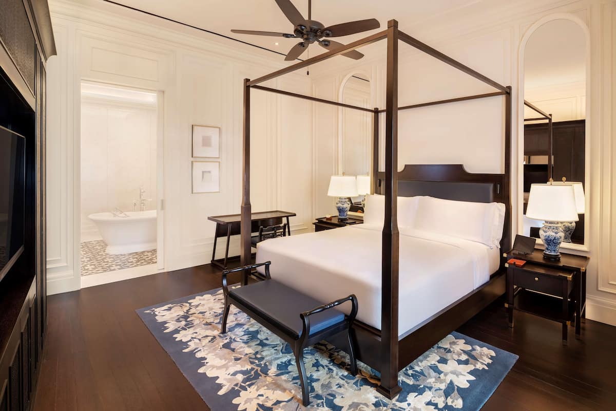 A four-poster bed with a wooden frame, a ceiling fan, a nightstand with a lamp, and a bathroom with a bathtub and sink.
