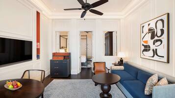 A modern living room with a blue sofa, a dining table with fruit, a ceiling fan, and a large abstract painting.