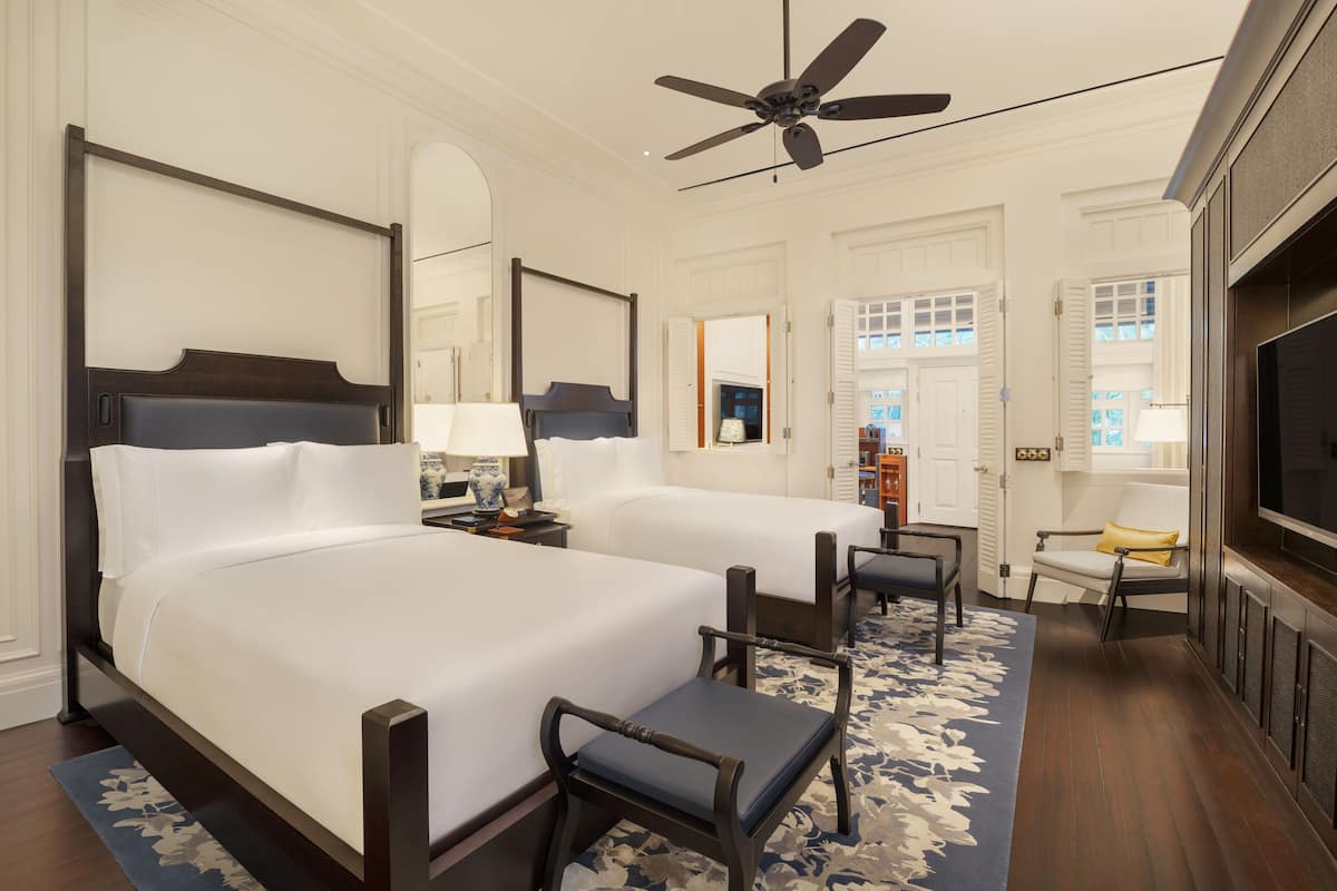 A hotel room with two beds, a ceiling fan, a TV, a chair, and a small table.
