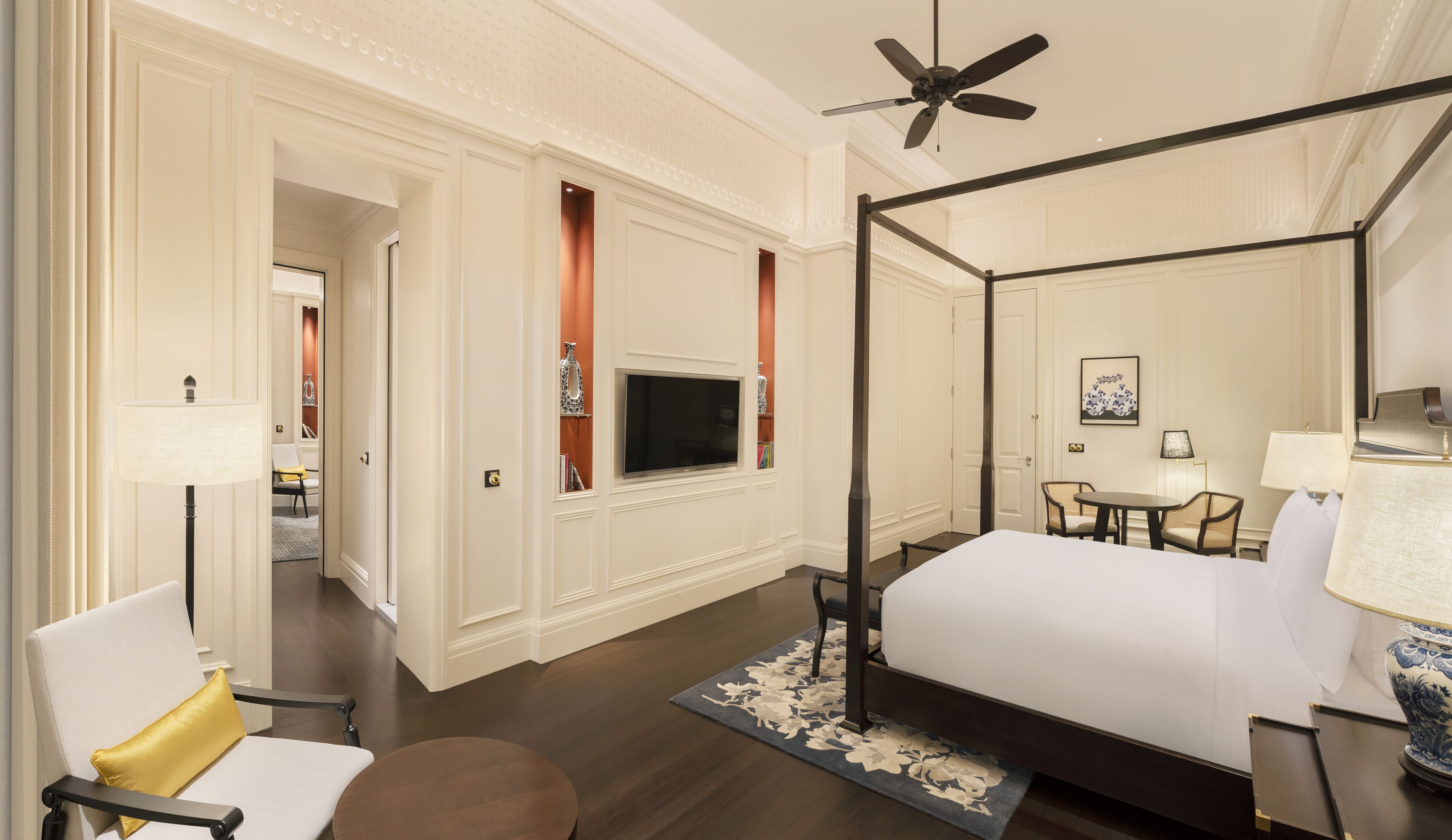 A hotel room with a four-poster bed, a television mounted on the wall, a dining area with a table and chairs, and a ceiling fan.