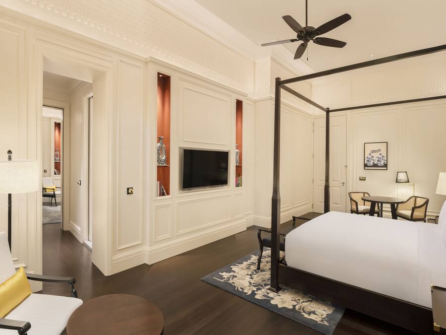 A hotel room with a four-poster bed, a television mounted on the wall, a dining area with a table and chairs, and a ceiling fan.