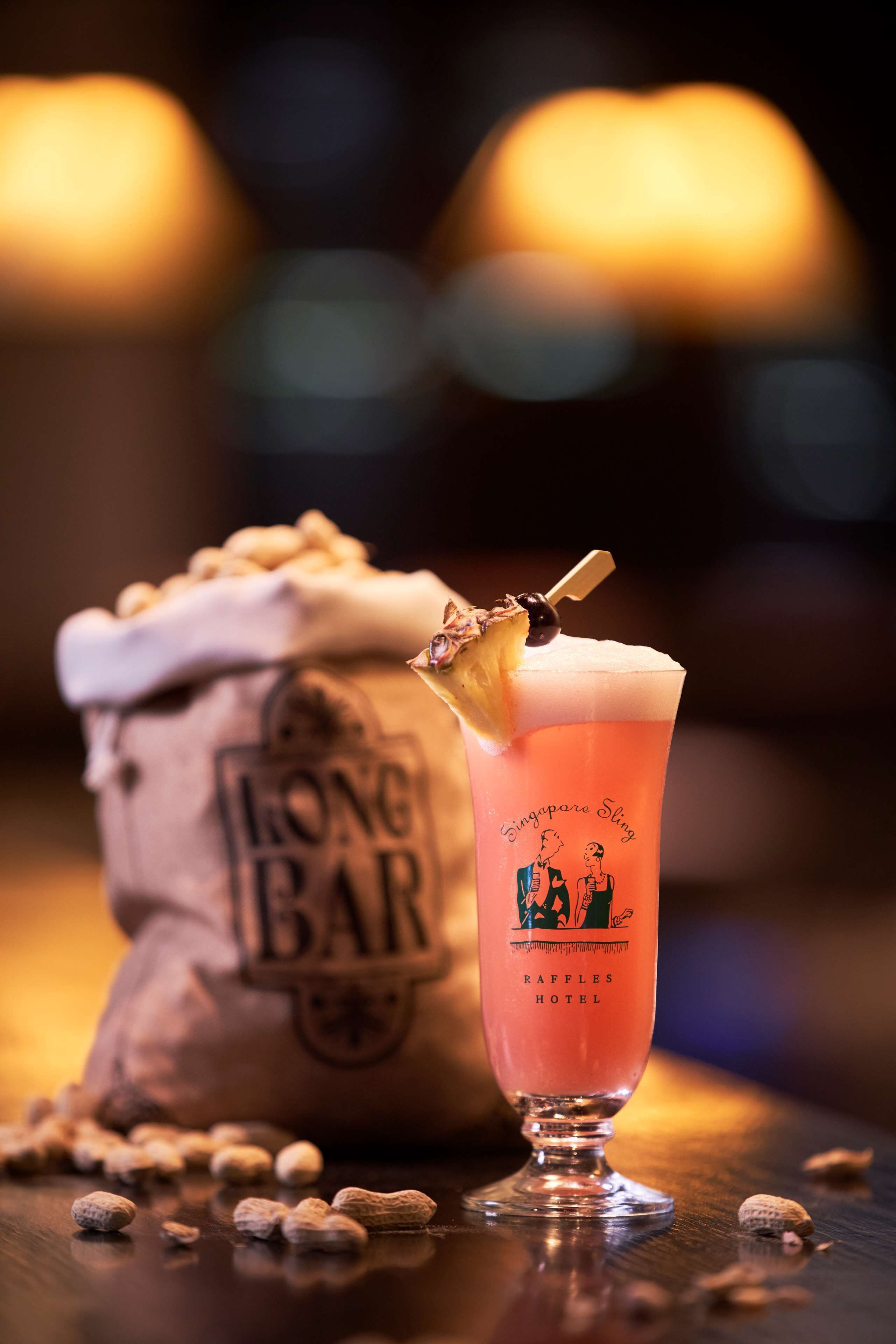 A cocktail glass with a garnish, a bag of peanuts, and a bag labeled "LONG BAR" on a wooden surface.