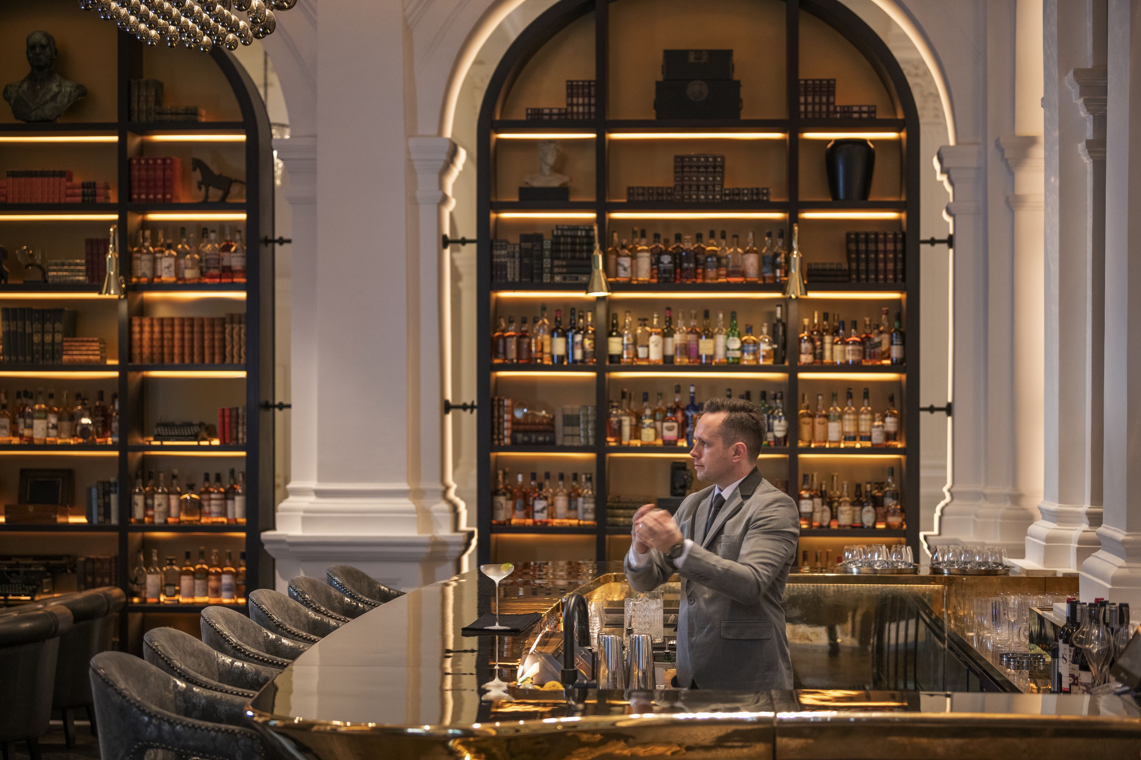 A bar area with shelves of liquor bottles, a bartender preparing drinks, and leather bar chairs.