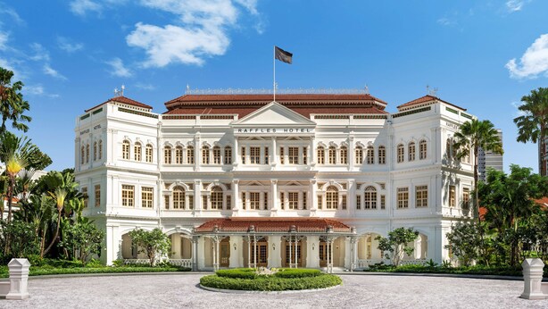 Un grand bâtiment blanc de style colonial, sur lequel on peut clairement lire l’inscription « RAFFLES HOTEL ». Il se caractérise par une façade symétrique, dotée de nombreuses fenêtres et d’une entrée centrale.