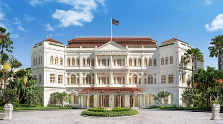 A large, white, colonial-style building with the name 'RAFFLES HOTEL' prominently displayed. It features a symmetrical facade with multiple windows and a central entrance.