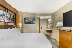 Habitación de hotel con una cama grande, un televisor de pantalla plana montado en la pared, un cuadro, un baño con lavamanos y espejo, y un armario de madera.