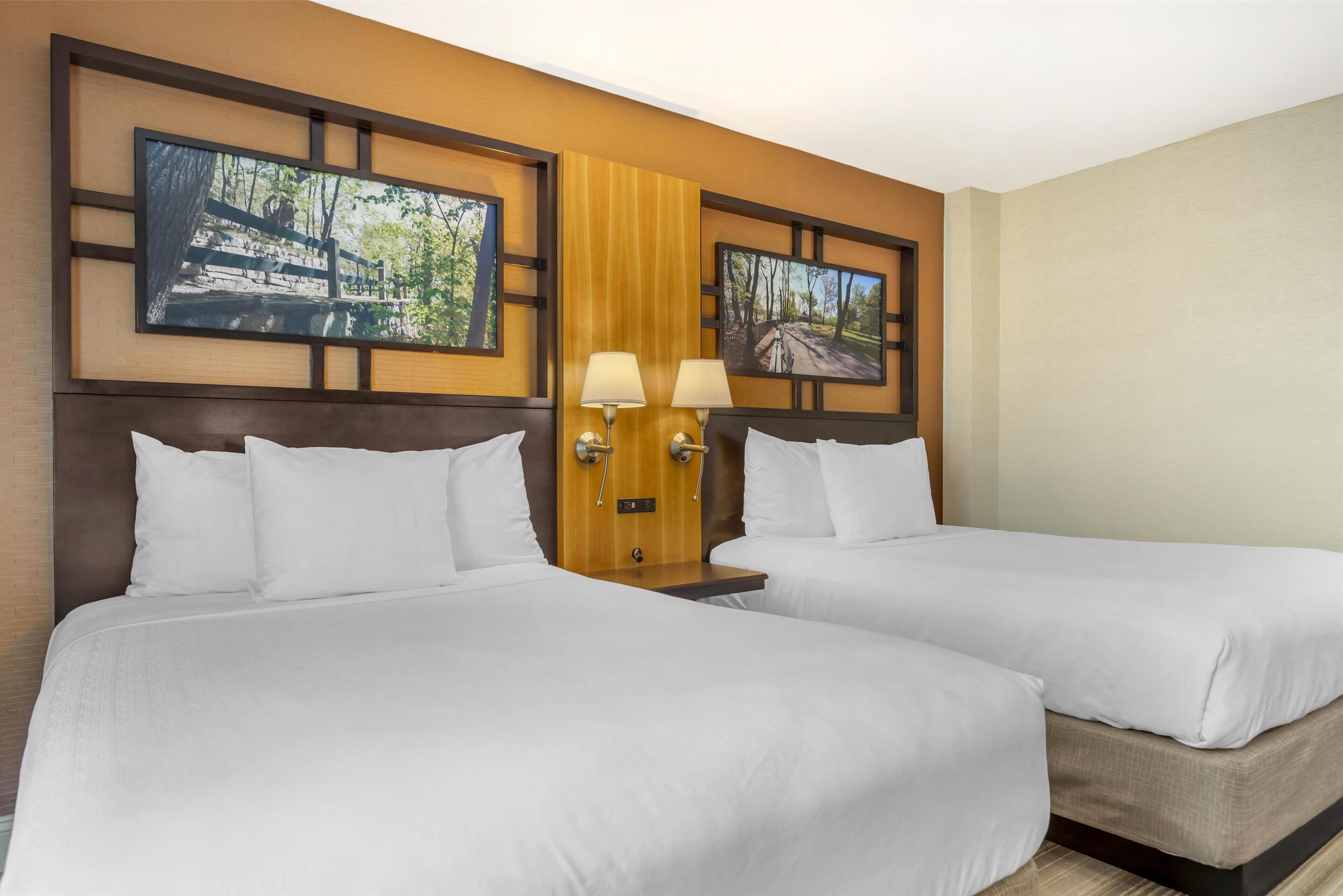 A hotel room with two beds, wooden headboard, two wall-mounted lamps, and two framed pictures on the wall.