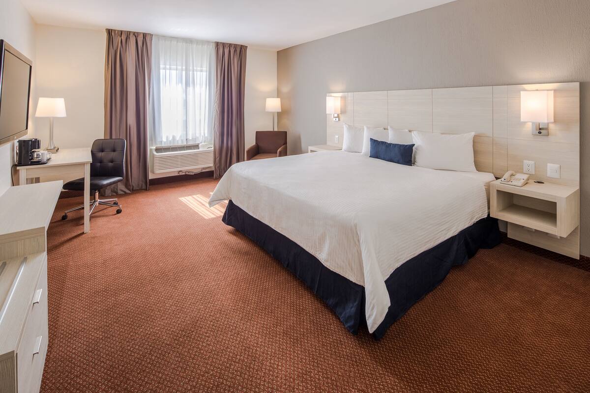 A hotel room with a large bed, a desk with a chair, a lamp, and a bedside table with a phone.
