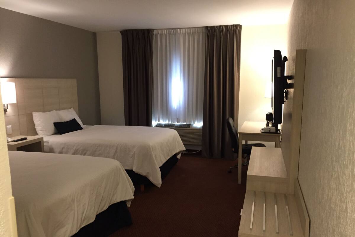 A hotel room with two beds, a desk, a chair, a lamp, a window with curtains, and an air conditioning unit.