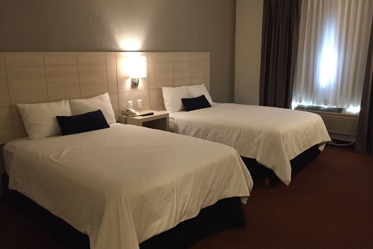 A hotel room with two beds, a nightstand with a phone, and a window with curtains.