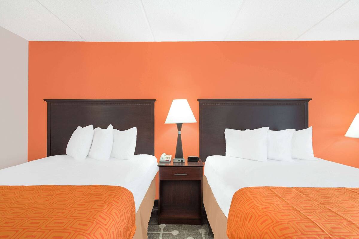 A hotel room with two beds, a nightstand with a lamp, and an orange accent wall.