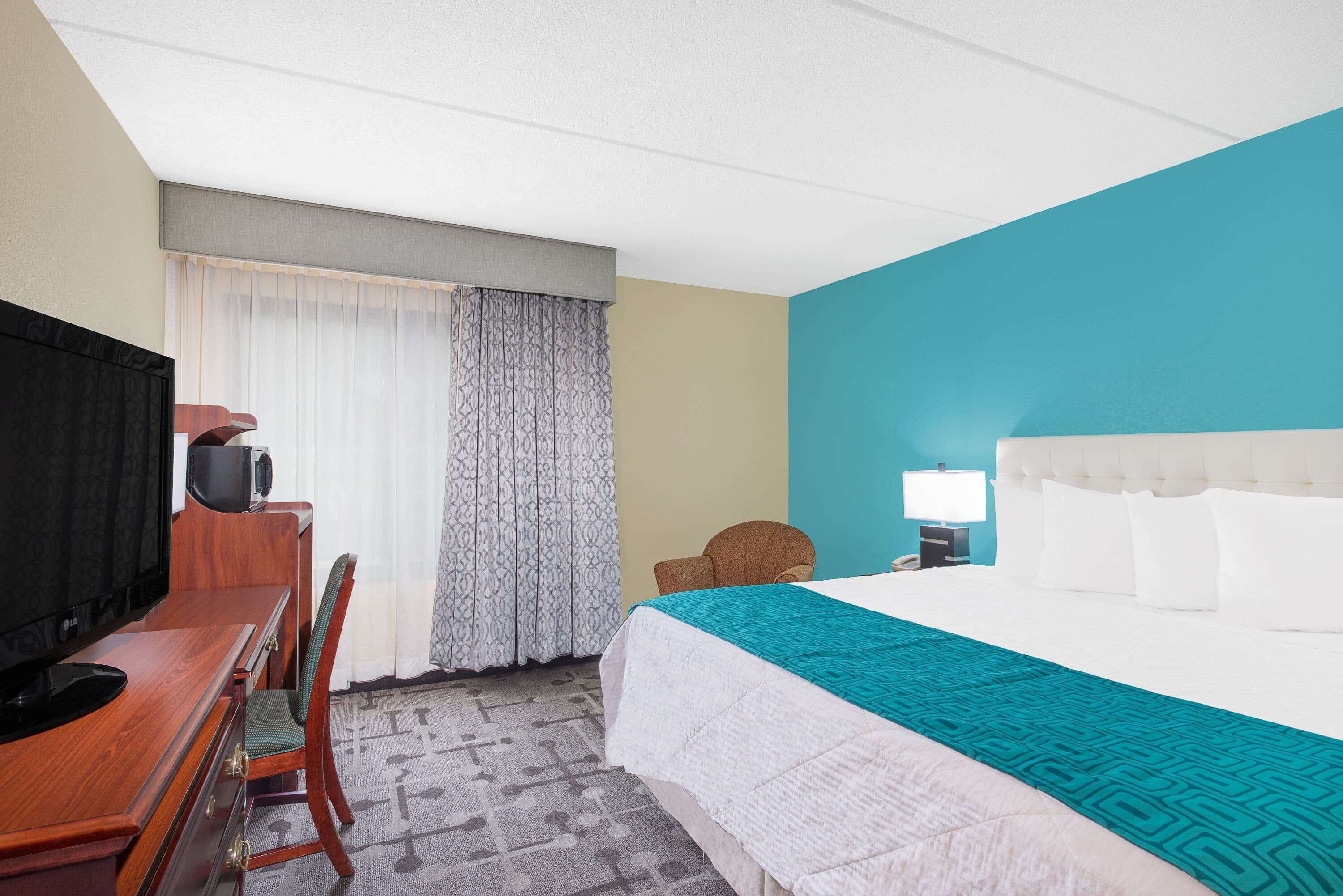 A hotel room with a bed, a desk with a television, a chair, and a window with curtains.