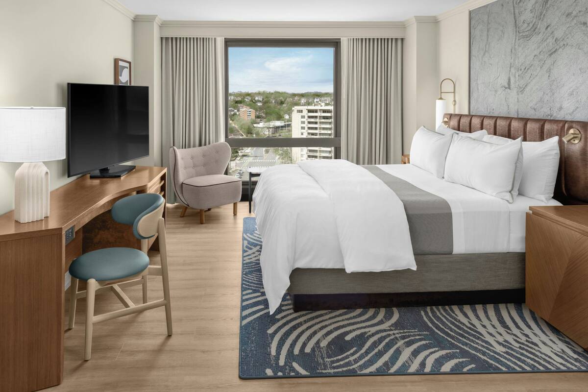 A hotel room with a bed, a desk with a TV, a chair, a sofa, and a view of buildings through the window.