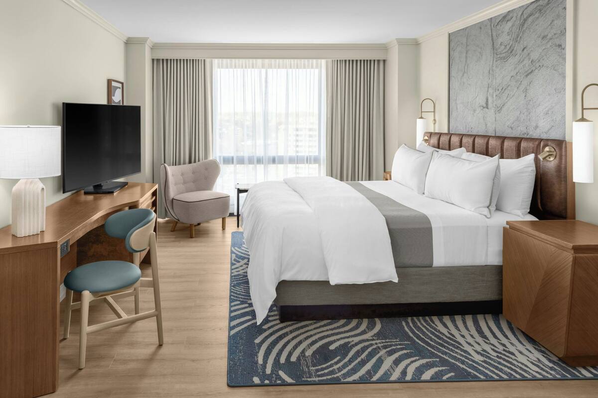 A hotel room with a bed, a desk with a television, a chair, and a view of the city through large windows.
