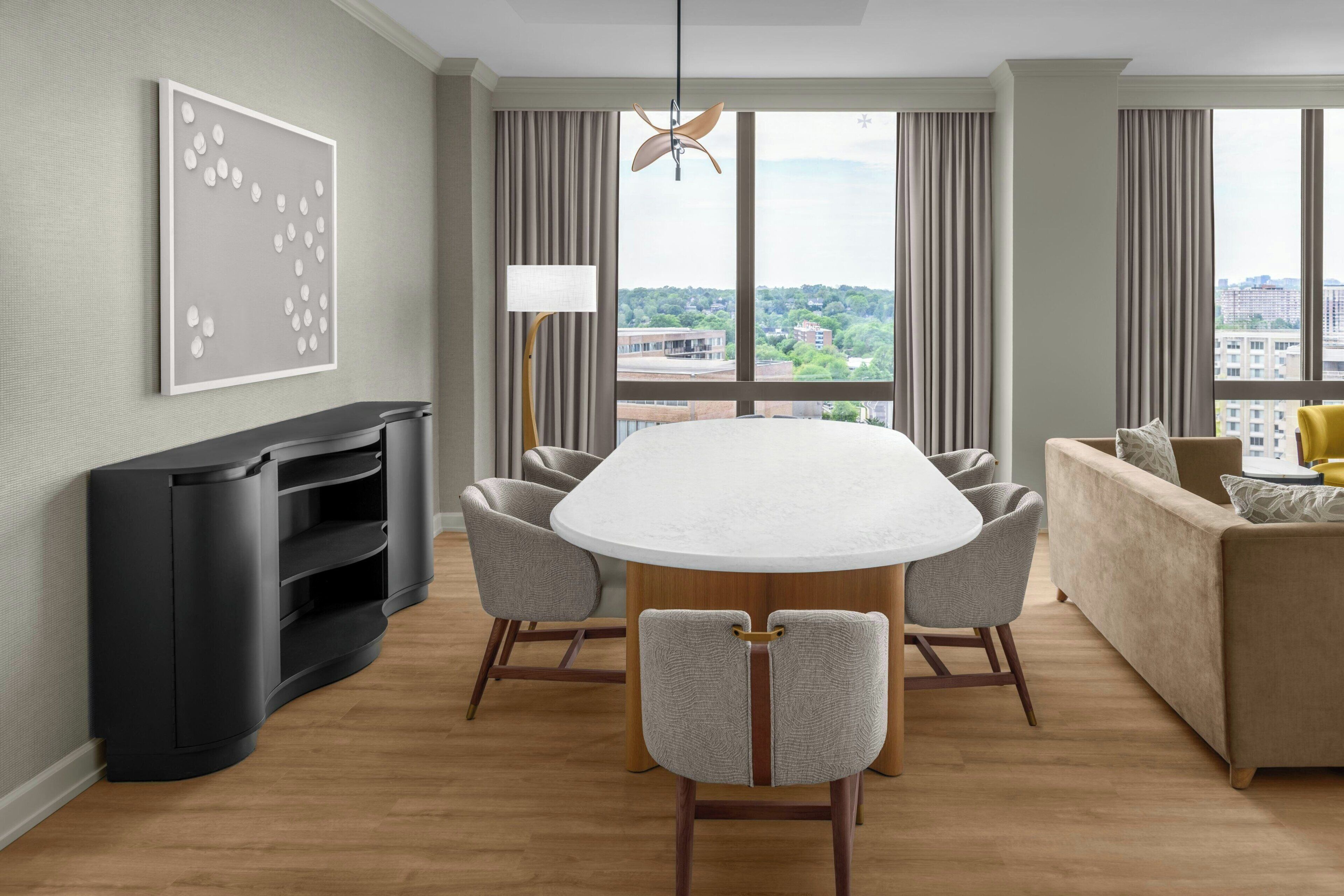 Una habitación de hotel moderna con una mesa de comedor, sillas, un sofá, un televisor y un amplio ventanal con vistas a la ciudad.