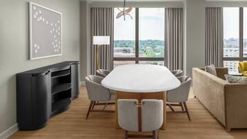 Une chambre d’hôtel moderne équipée d’une table à manger, de chaises, d’un canapé, d’une télévision et d’une grande fenêtre donnant sur la ville.
