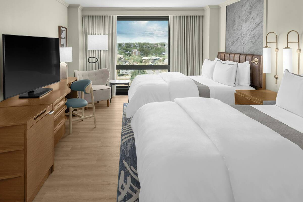 A hotel room with two beds, a desk with a TV, a chair, and a view of the city.