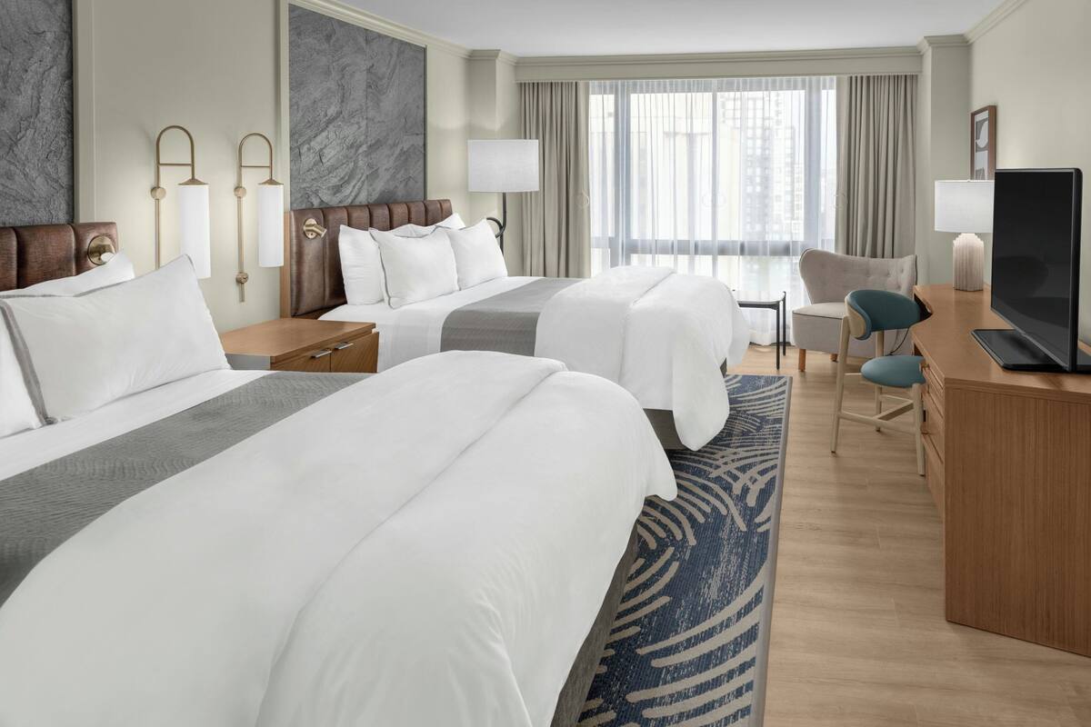 A hotel room with two beds, a desk, a chair, a TV, and a large window with curtains.