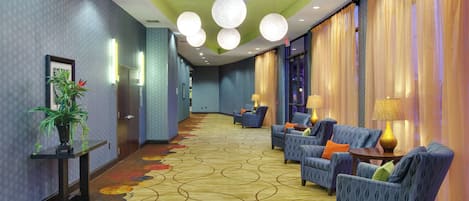 A hotel corridor with a patterned carpet, seating area with armchairs, and a reception desk with a plant.
