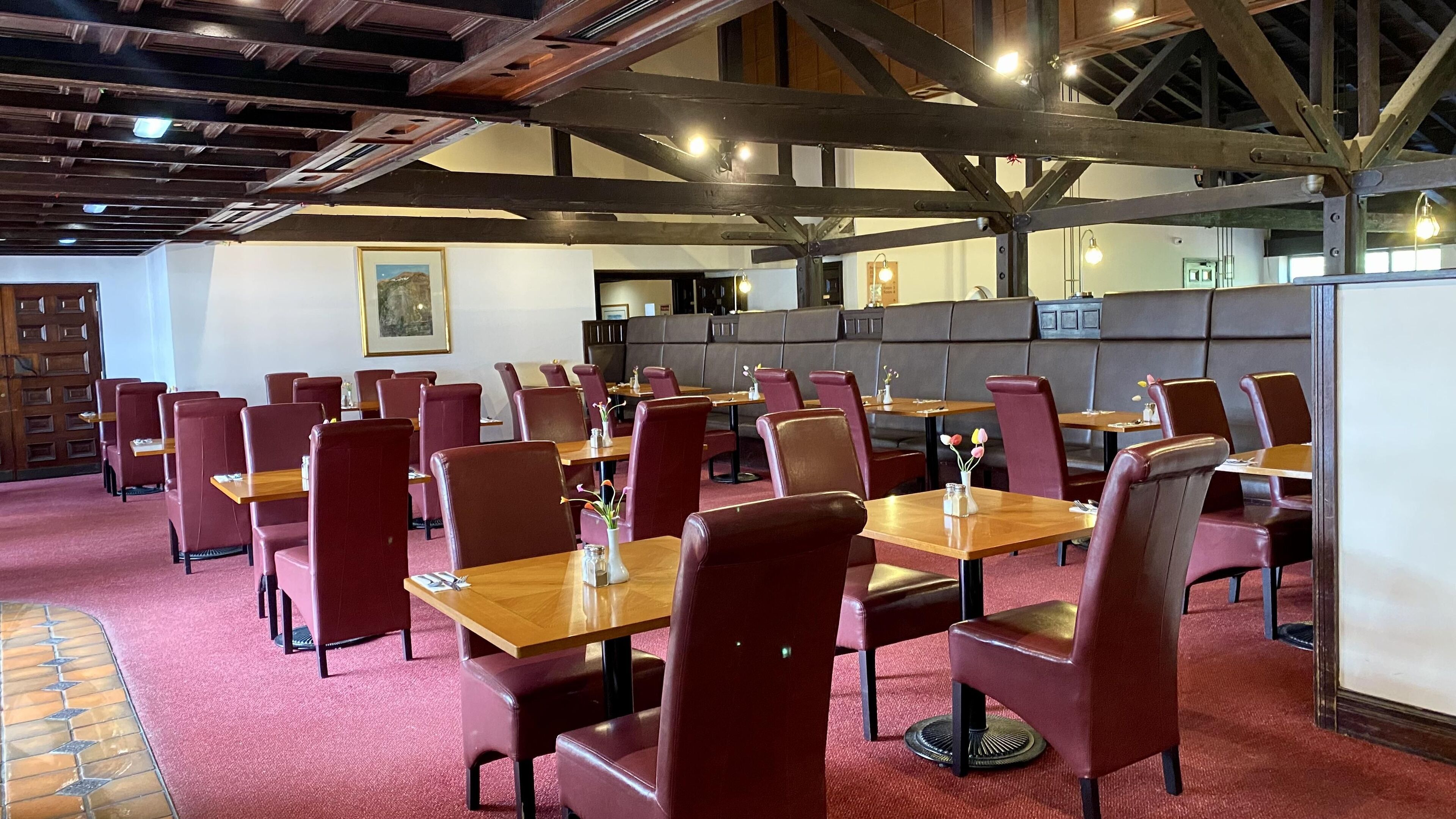 Un restaurant avec des tables en bois et des chaises rouges, un plafond haut avec des poutres apparentes et un sol recouvert d’une moquette rouge.