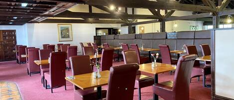 Un restaurant avec des tables en bois et des chaises rouges, un plafond haut avec des poutres apparentes et un sol recouvert d’une moquette rouge.