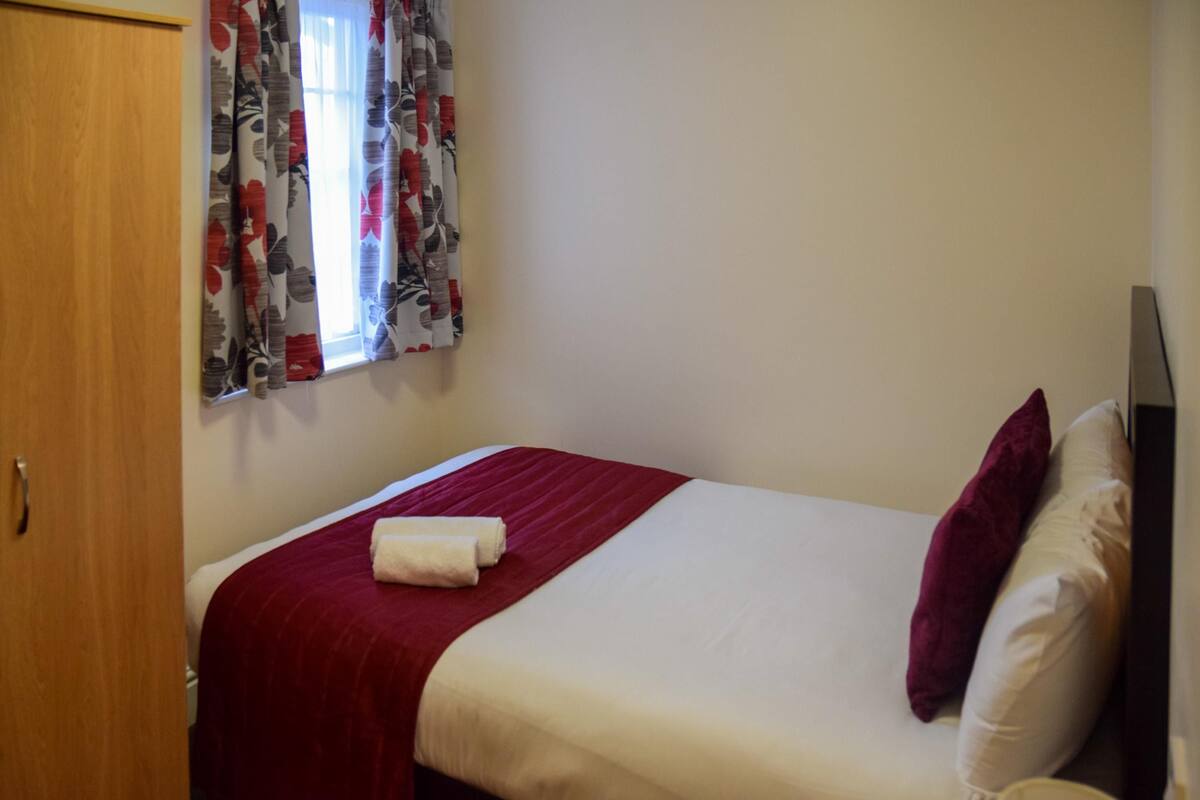 A single bed with a red blanket and white pillows, a wooden wardrobe, and a window with floral curtains.