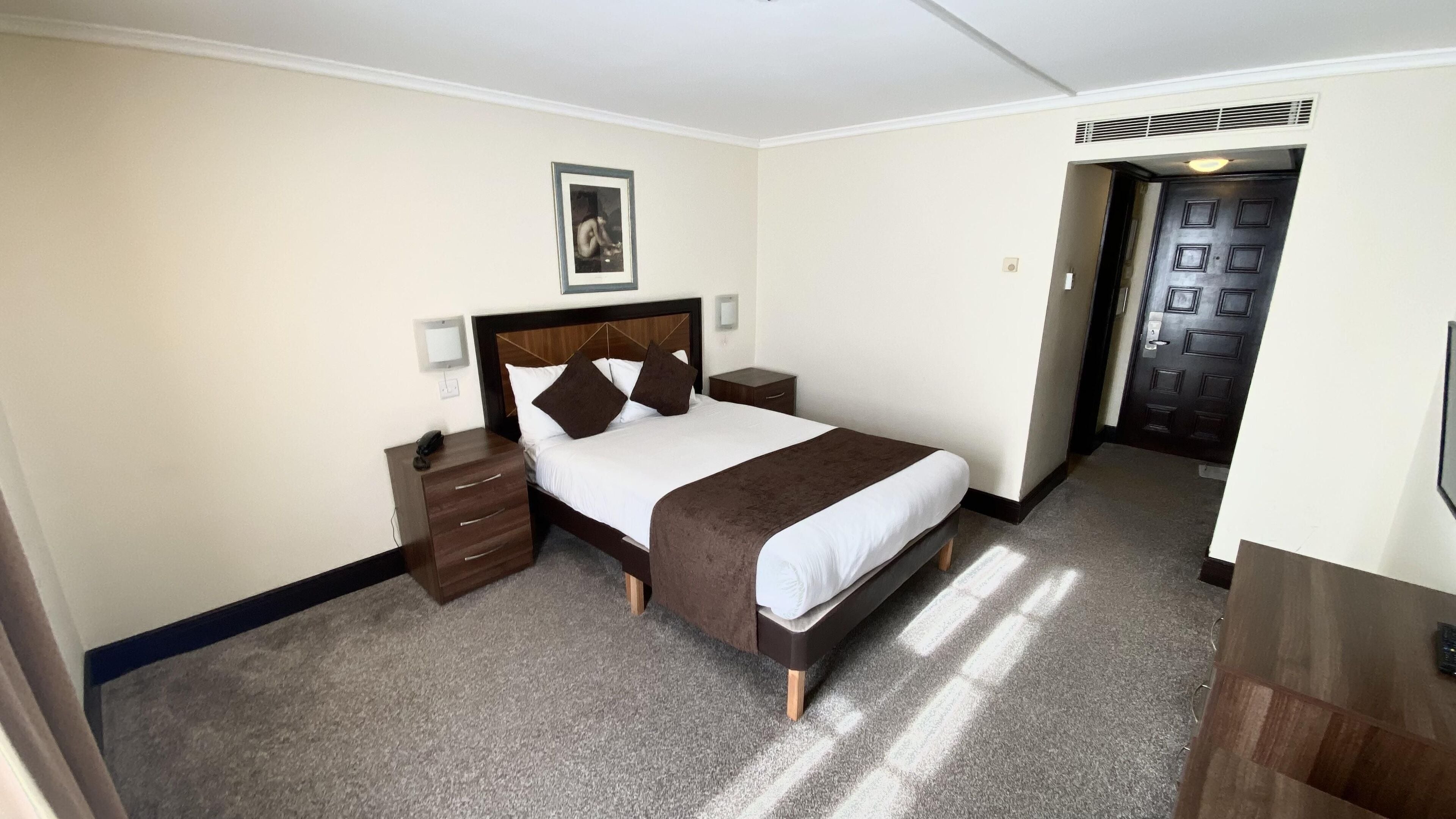 Habitación de hotel con cama, mesitas de noche, un cuadro enmarcado en la pared y una puerta con respiradero.