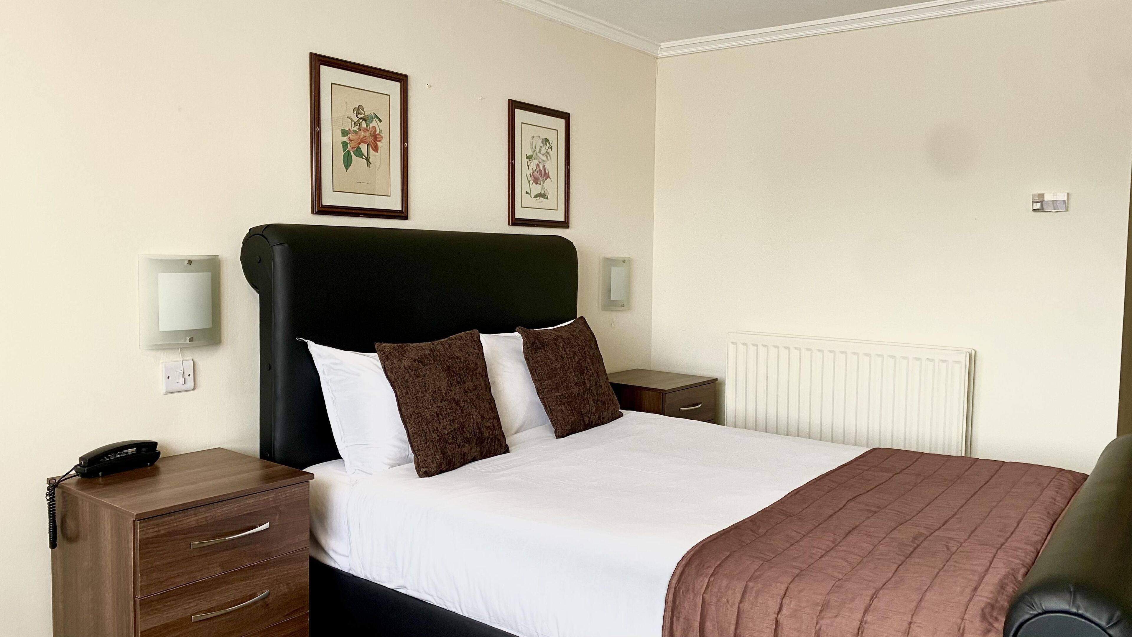 A neatly made bed with white and brown bedding, a black headboard, and two framed botanical prints on the wall.