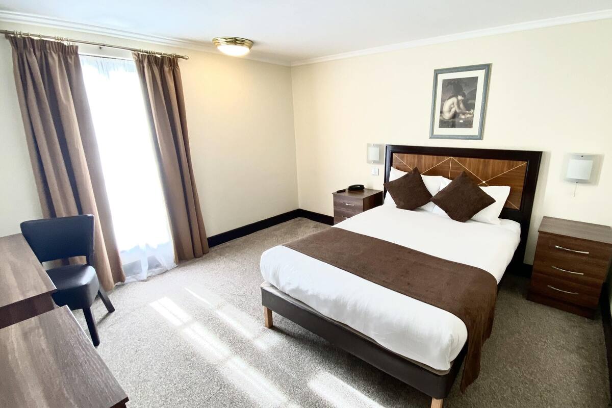 A hotel room with a large bed, a desk, a chair, a nightstand, and a picture on the wall.