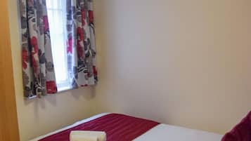Una cama individual con una colcha roja y un almohadón blanco, una mesita de noche de madera con un control remoto y una lámpara, y una ventana con cortinas estampadas.
