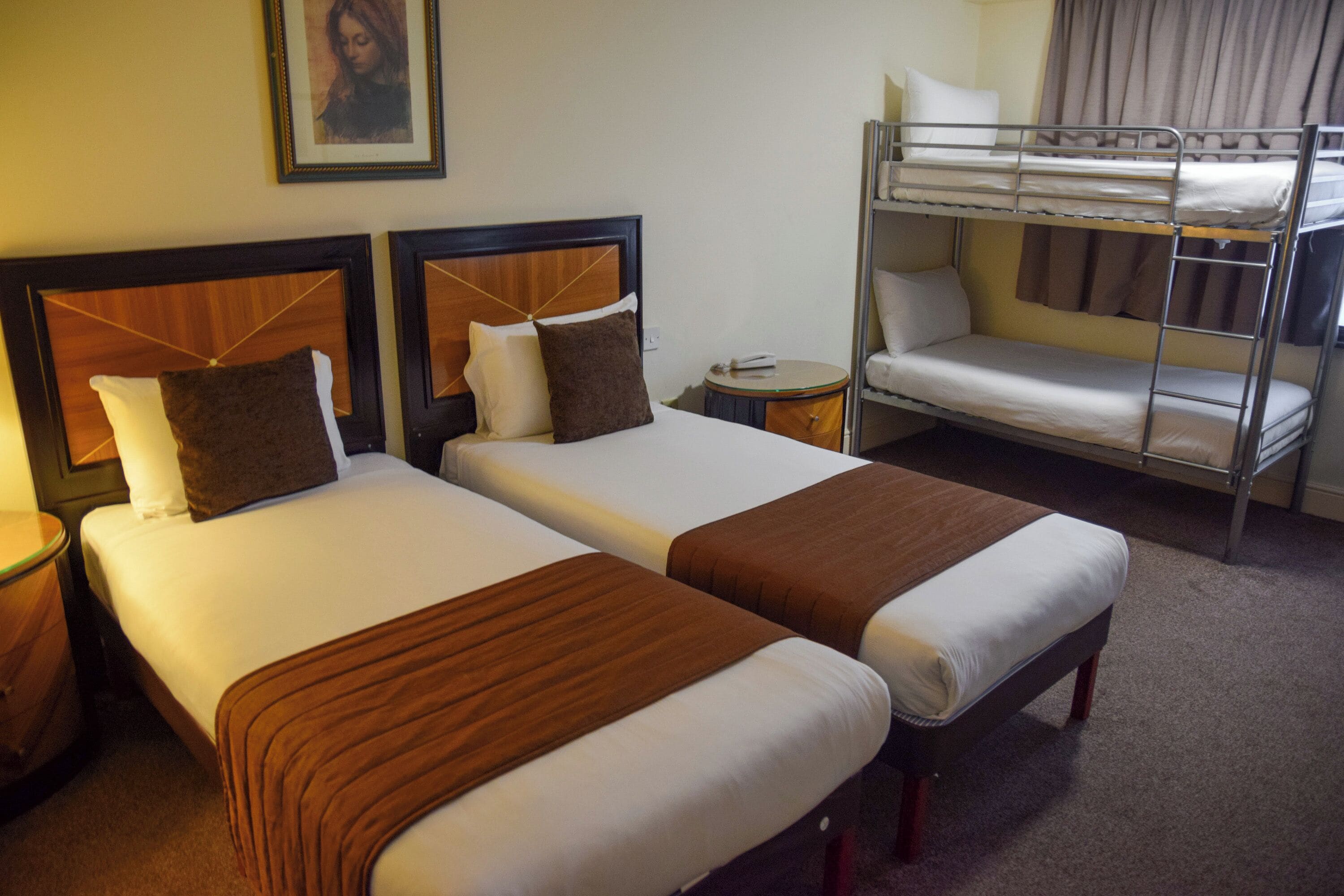 A hotel room with two single beds, a bunk bed, a bedside table, and a framed picture on the wall.