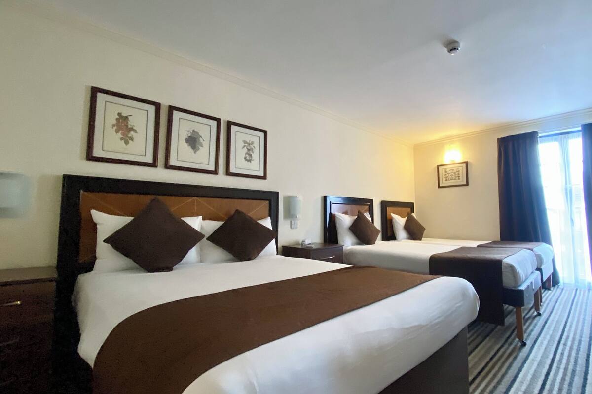 A hotel room with two beds, a desk, and a chair. There are framed pictures on the wall and a window with curtains.