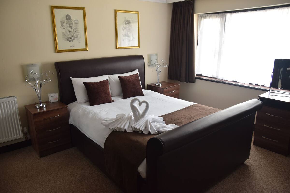 A hotel room with a large bed, two bedside tables, a nightstand, a dresser, a window with curtains, and two framed pictures on the wall.