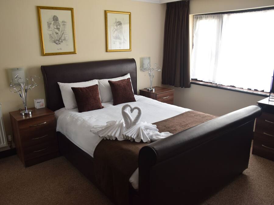 A hotel room with a large bed, two bedside tables, a nightstand, a dresser, a window with curtains, and two framed pictures on the wall.
