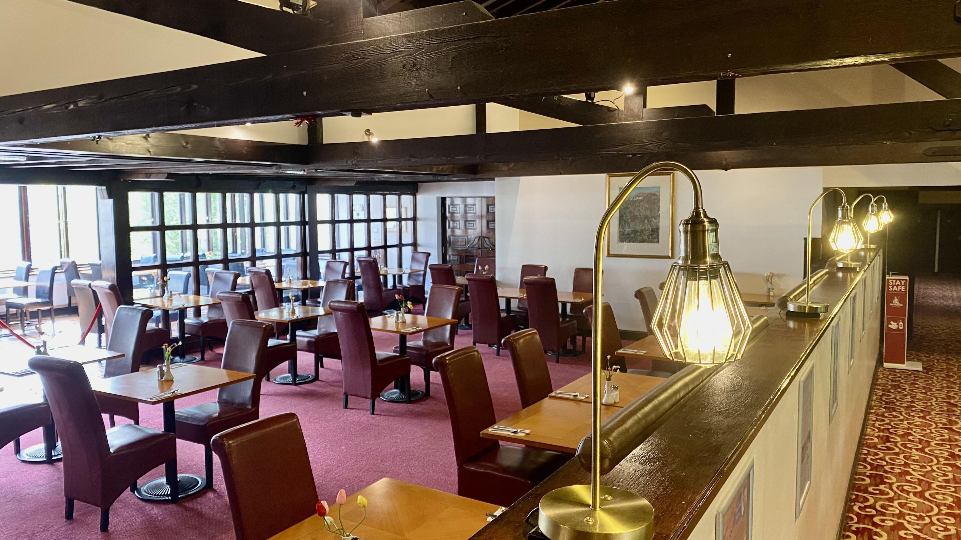Un restaurante con techos altos, vigas a la vista y una larga barra con una lámpara de estilo antiguo. Las mesas están dispuestas para comer y hay grandes ventanales que dejan entrar luz natural.