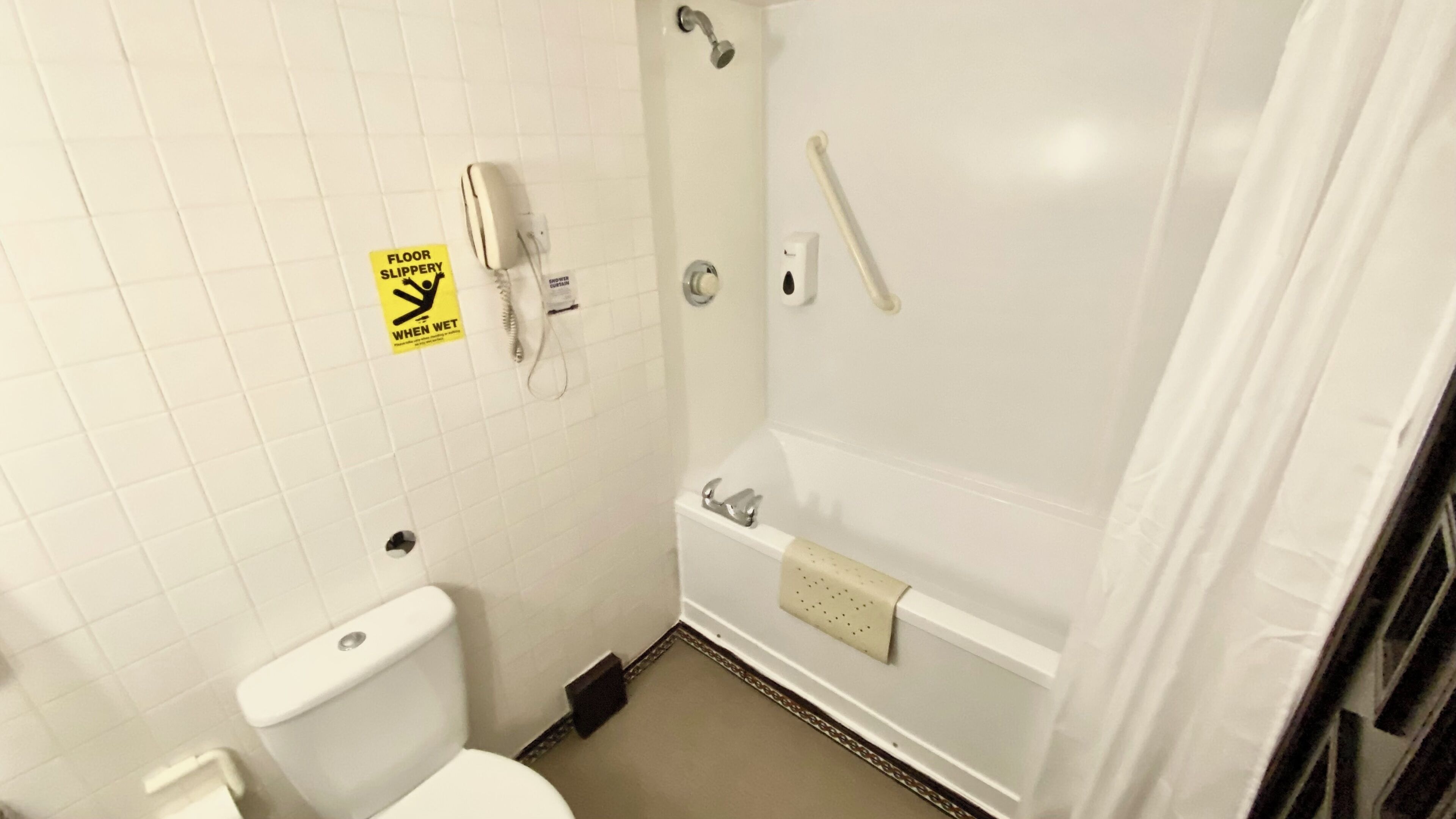 Un baño con inodoro, ducha con barra de apoyo y letrero que advierte sobre suelo resbaladizo cuando está mojado.