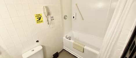 Un baño con inodoro, ducha con barra de apoyo y letrero que advierte sobre suelo resbaladizo cuando está mojado.