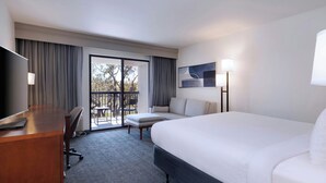 Ein Hotelzimmer mit einem großen Bett, einem Schreibtisch mit Stuhl, einer Couch, einem Fernseher und einem Balkon mit Blick auf Bäume.