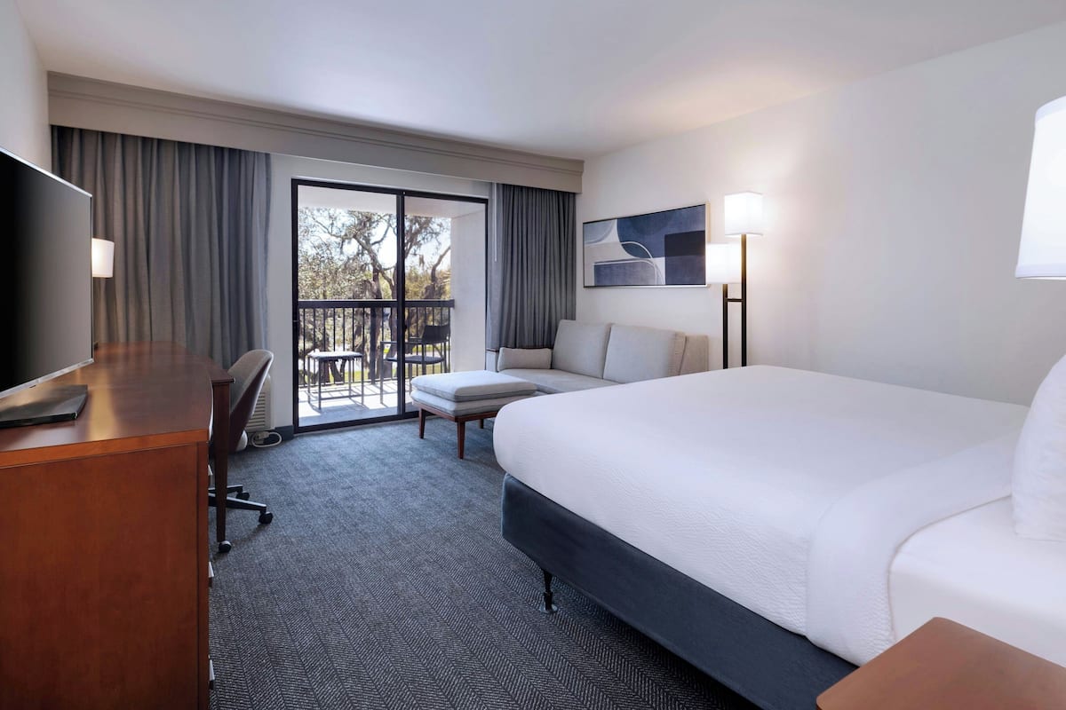 Une chambre d’hôtel avec un grand lit, un bureau avec une chaise, un canapé, une télévision et un balcon donnant sur la verdure.