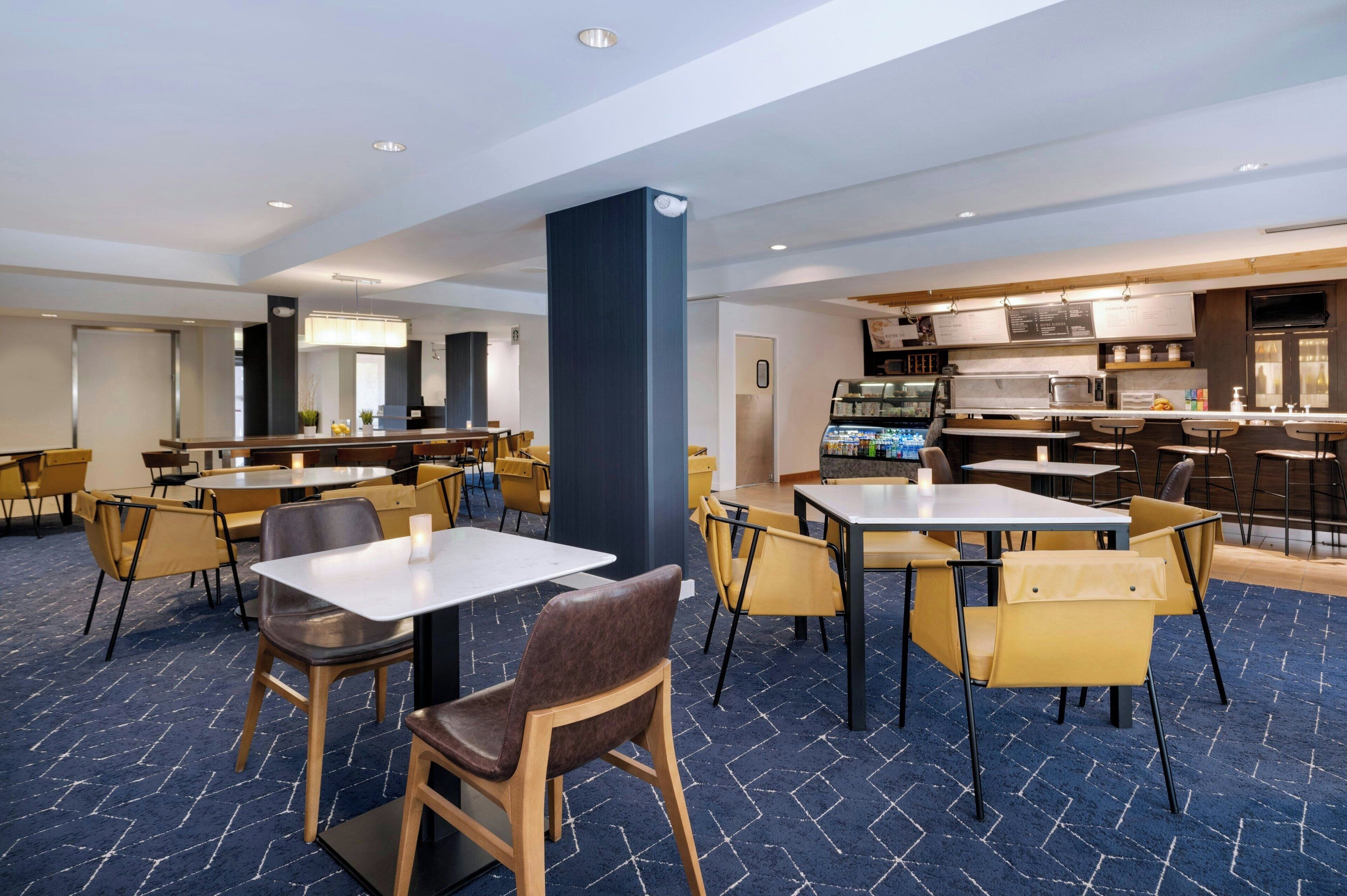A modern hotel dining area with multiple tables and chairs, a central pillar, and a kitchen area in the background.
