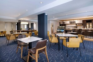 A modern hotel dining area with multiple tables and chairs, a central pillar, and a kitchen area in the background.