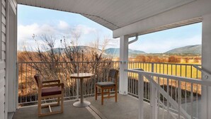 Un balcon abrité, doté de deux chaises en bois et d’une petite table ronde, offrant une vue panoramique sur un paysage magnifique, avec des montagnes et des champs.
