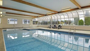 Une piscine intérieure à l’eau d’un bleu cristallin, avec des poutres en bois au plafond et une vue sur les arbres à travers de grandes fenêtres.