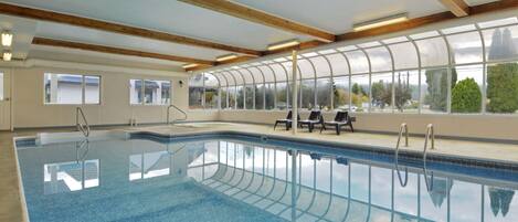 An indoor swimming pool with clear blue water, wooden beams on the ceiling, and a view of trees through large windows.
