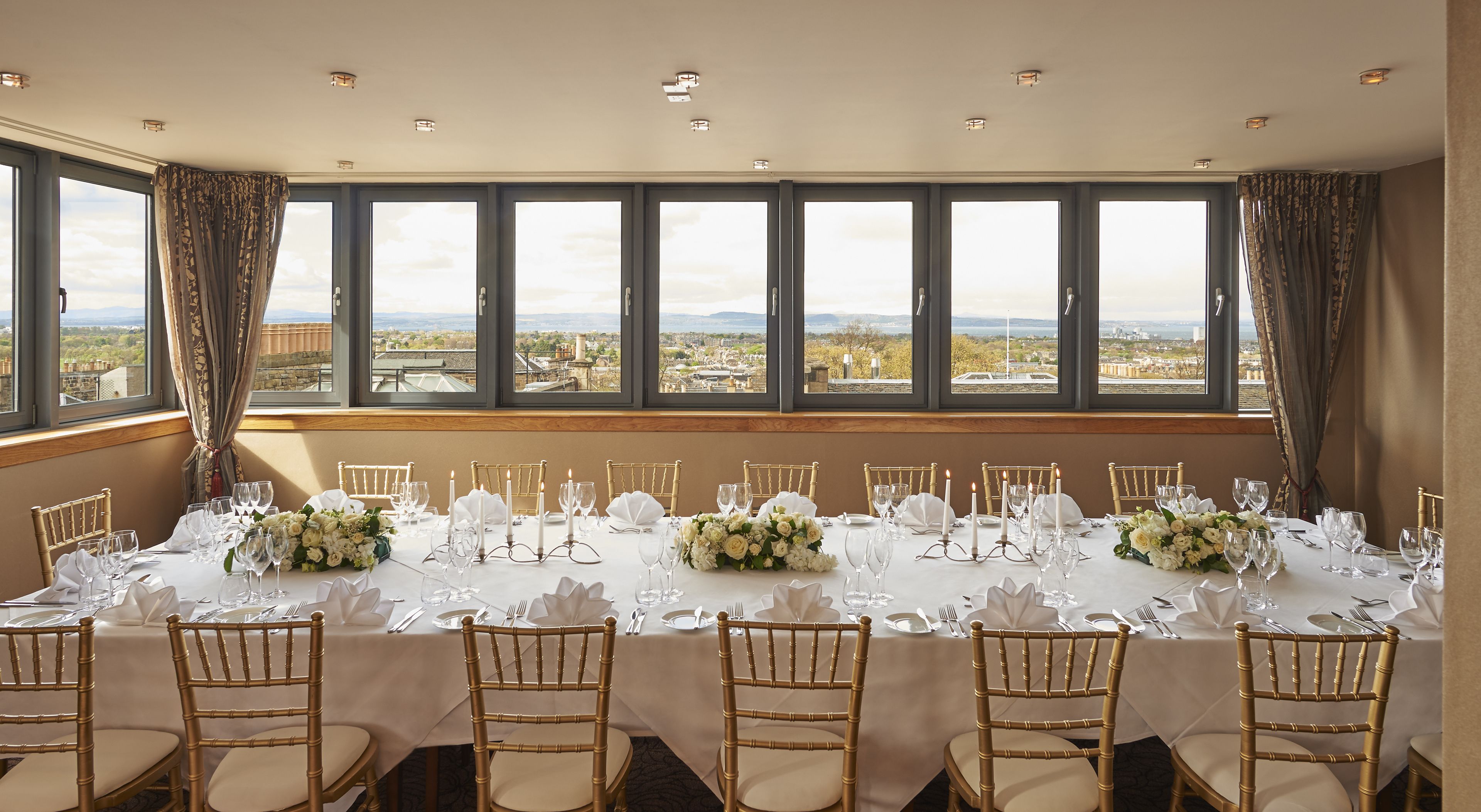 Una mesa de banquete dispuesta para un evento formal, con vista a un paisaje urbano a través de amplios ventanales.