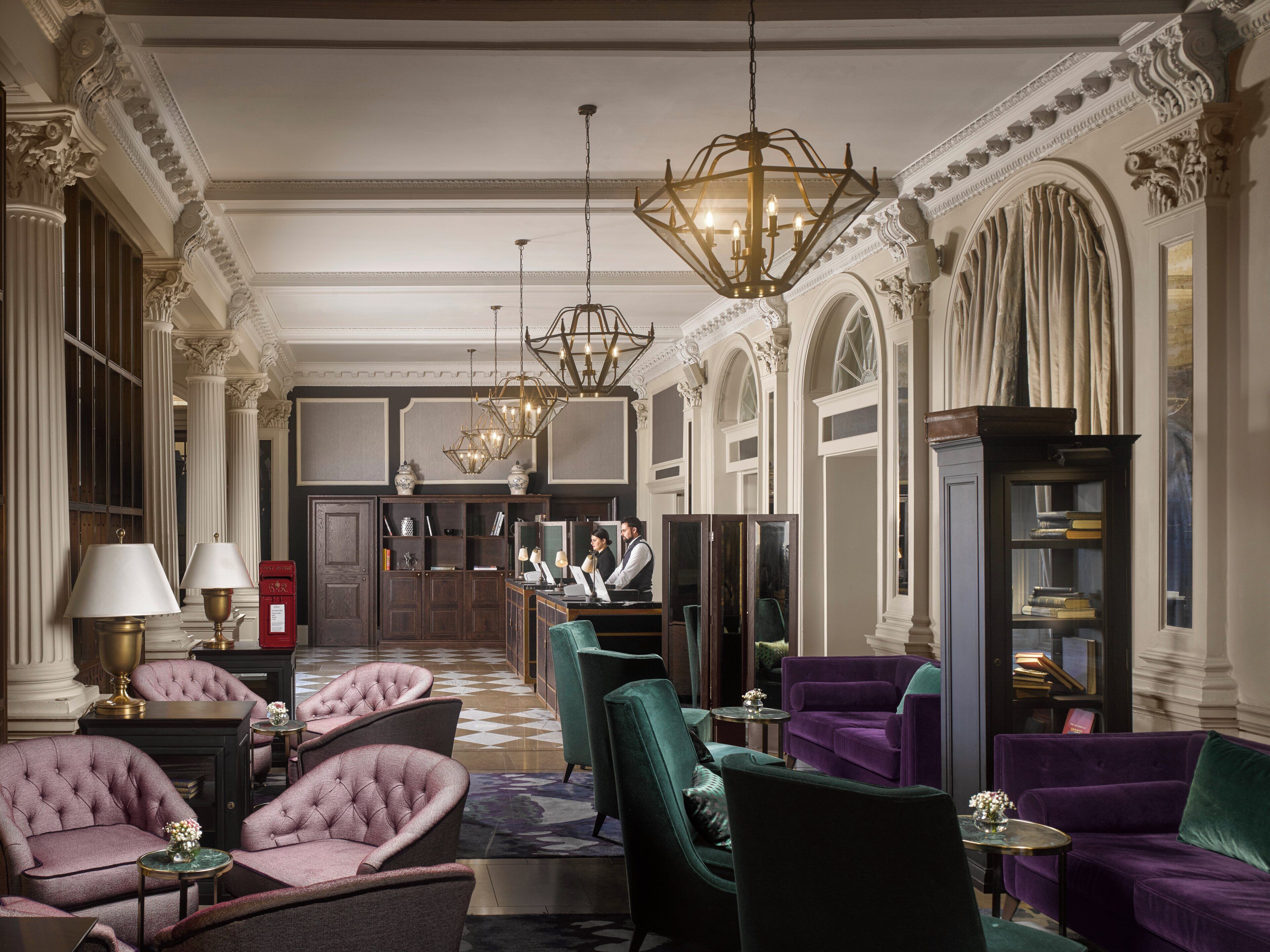 A spacious lobby with high ceilings, ornate columns, and a grand chandelier. There are plush seating areas and a reception desk in the background.